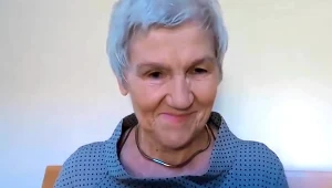 Starsza kobieta z krótkimi, siwymi włosami uśmiecha się lekko, ubrana w szarą bluzkę w drobne kropki, na szyi ma srebrny naszyjnik, siedzi na drewnianym krześle przy jasnym tle.