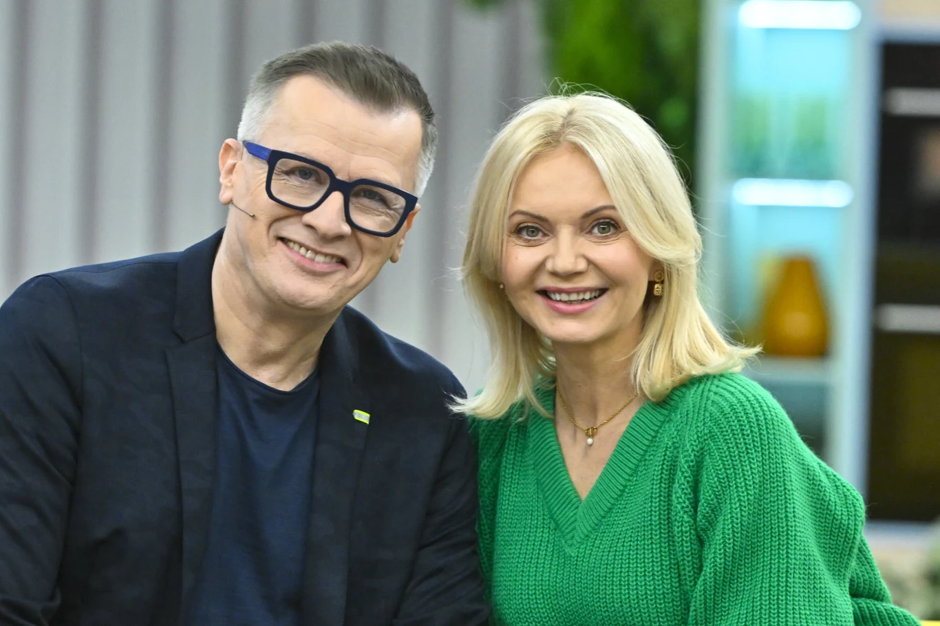 Uśmiechnięta para dorosłych ubrana w casualowe stroje, mężczyzna w okularach i ciemnej marynarce oraz kobieta w zielonym swetrze, siedzą blisko siebie na tle nowoczesnego, jasnego wnętrza.