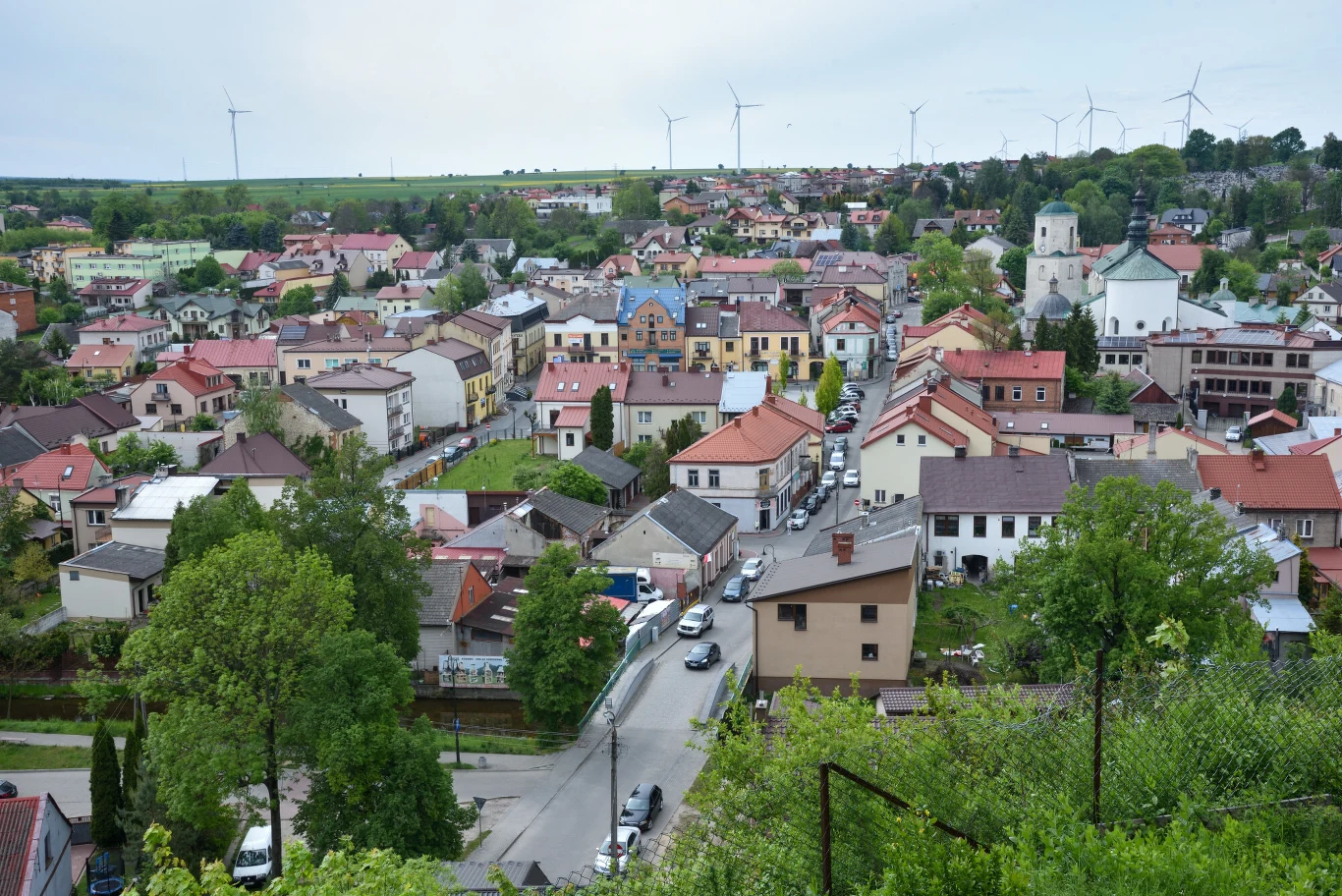 Niska zabudowa mieszkalna z czerwonymi dachami, w centrum widoczny charakterystyczny budynek z niebieskimi elementami, po prawej stronie kościół z jasną wieżą, w tle linia nowoczesnych turbin wiatrowych na zielonych polach.