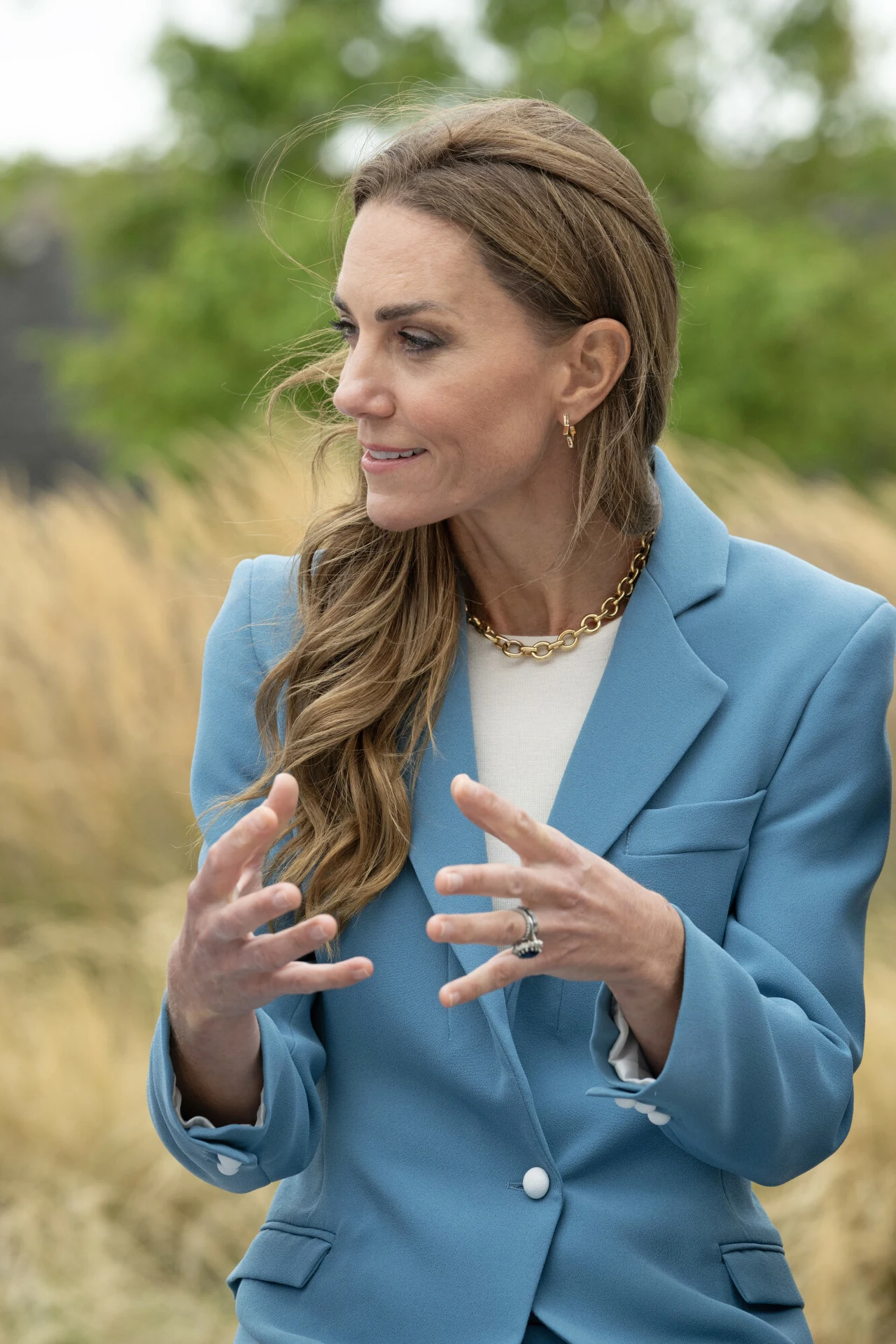 Mały detal w stylizacji Kate miał ogromne znaczenie Kobieta w niebieskiej marynarce i białej bluzce gestykuluje rękami, stojąc na tle wysokiej trawy i zieleni.