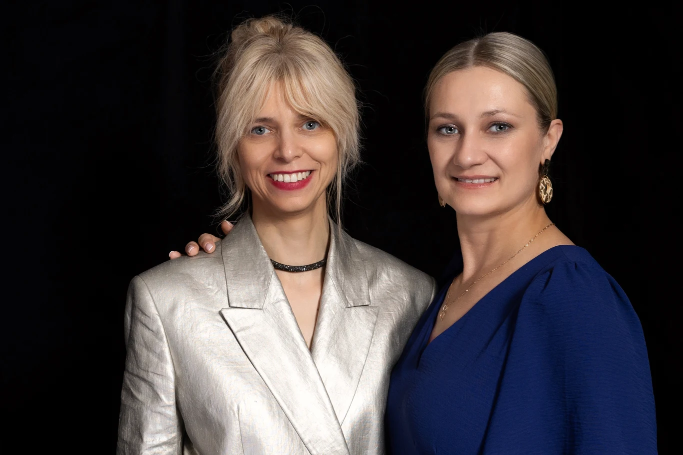 Dwie uśmiechnięte kobiety pozują razem na czarnym tle, jedna ubrana w srebrzystą marynarkę, druga w granatową sukienkę z odkrytym dekoltem i złotymi kolczykami.