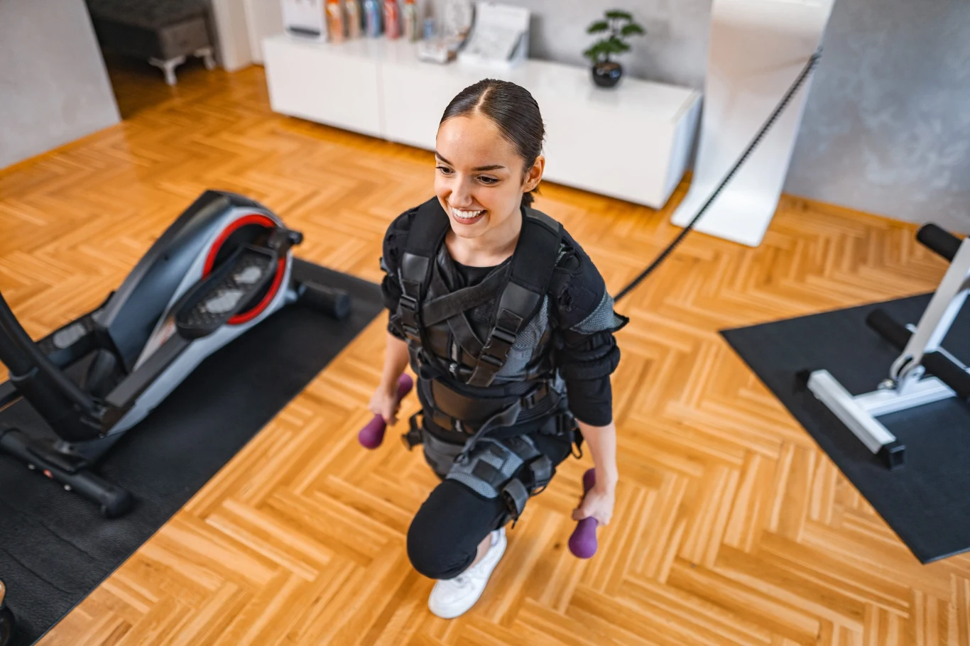 Trening EMS kusi krótkim czasem i obietnicą szybkich efektów. Czy 20 minut z elektrostymulacją naprawdę może zastąpić klasyczny trening? Kobieta w kombinezonie EMS ćwiczy z hantlami na siłowni, wykorzystując elektrostymulację mięśni.