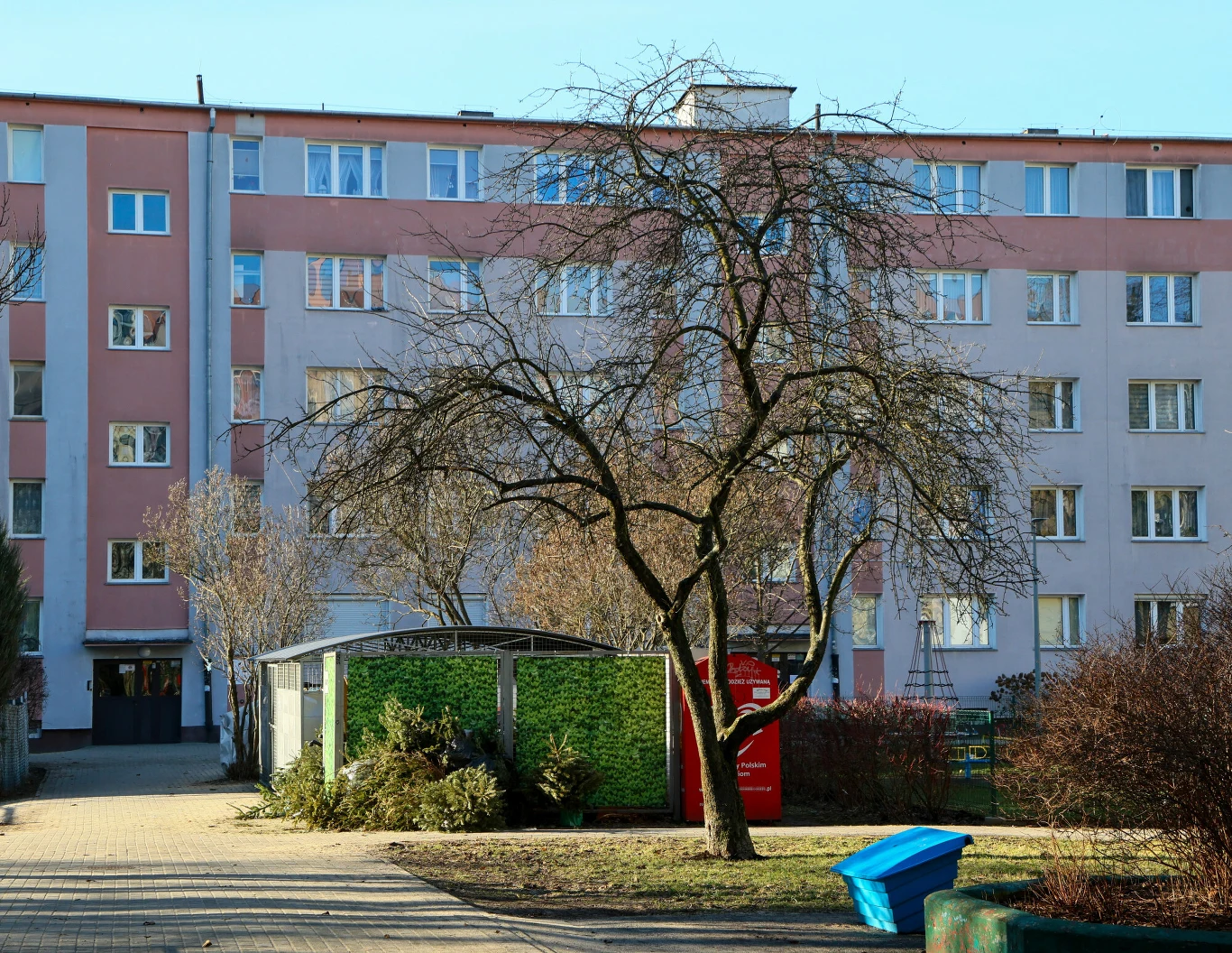 Osób, szukających perełek w altankach śmietnikowych i na wystawkach nie brakuje Blok mieszkalny w pastelowych kolorach z widocznym bezlistnym drzewem na pierwszym planie, wiata śmietnikowa oraz czerwona budka telefoniczna po prawej stronie drzewa.
