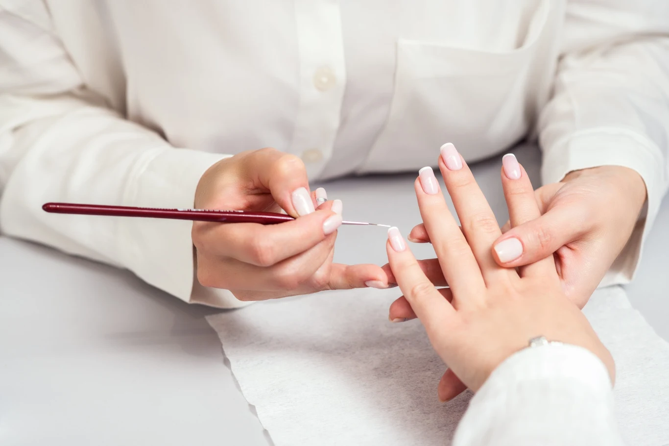 Jaki manicure będzie modny jesienią? Jest subtelny i elegancki Manicurzystka maluje jasnym lakierem paznokcie klientki trzymane na białym ręczniku.