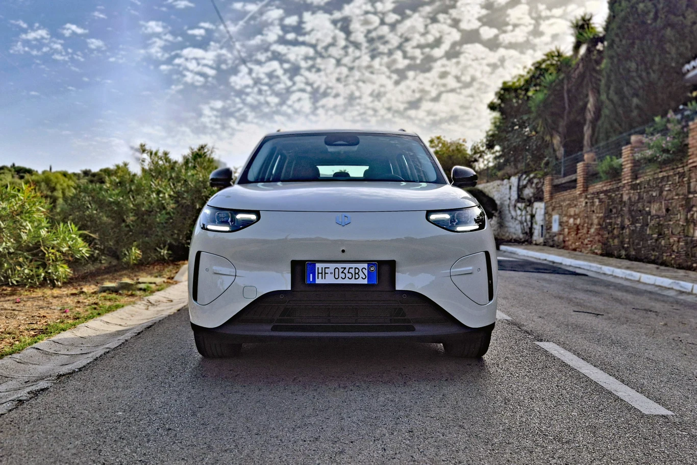 Leapmotor B03X Biały samochód elektryczny zaparkowany na środku asfaltowej drogi, otoczony zielenią i kamiennym murem po jednej stronie.