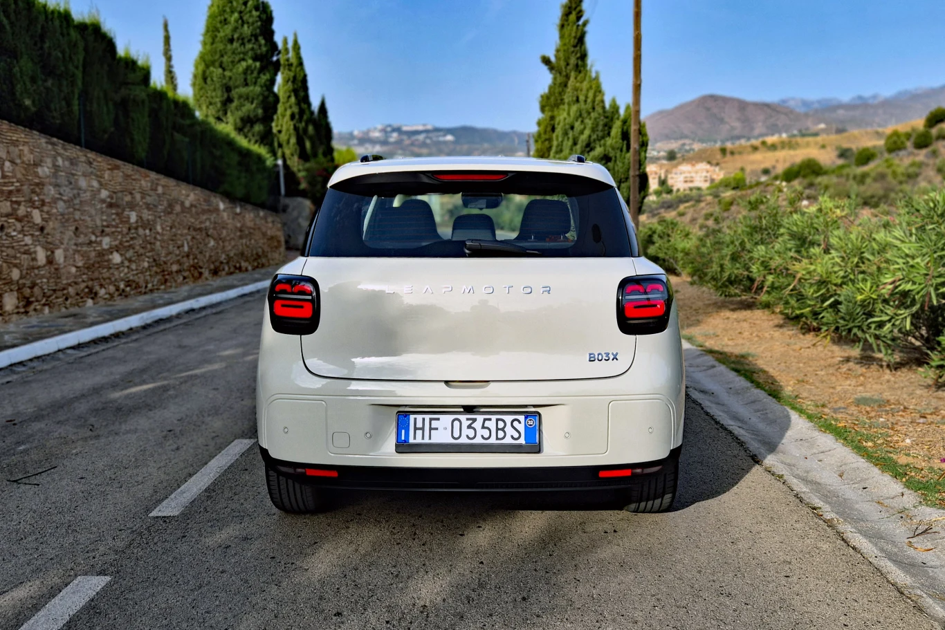 Leapmotor B03X Białe auto osobowe marki BAOJUN ustawione na środku wąskiej, asfaltowej drogi, otoczonej kamiennym murem i zielonymi drzewami, z widokiem na górzysty krajobraz w tle.