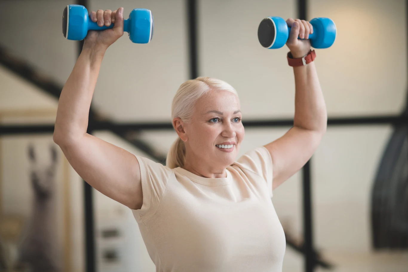 Trening z obciążeniem znacząco wzmacnia kości - potwierdzają badania Uśmiechnięta kobieta z blond włosami unosi nad głową niebieskie hantle, ćwicząc ramiona.