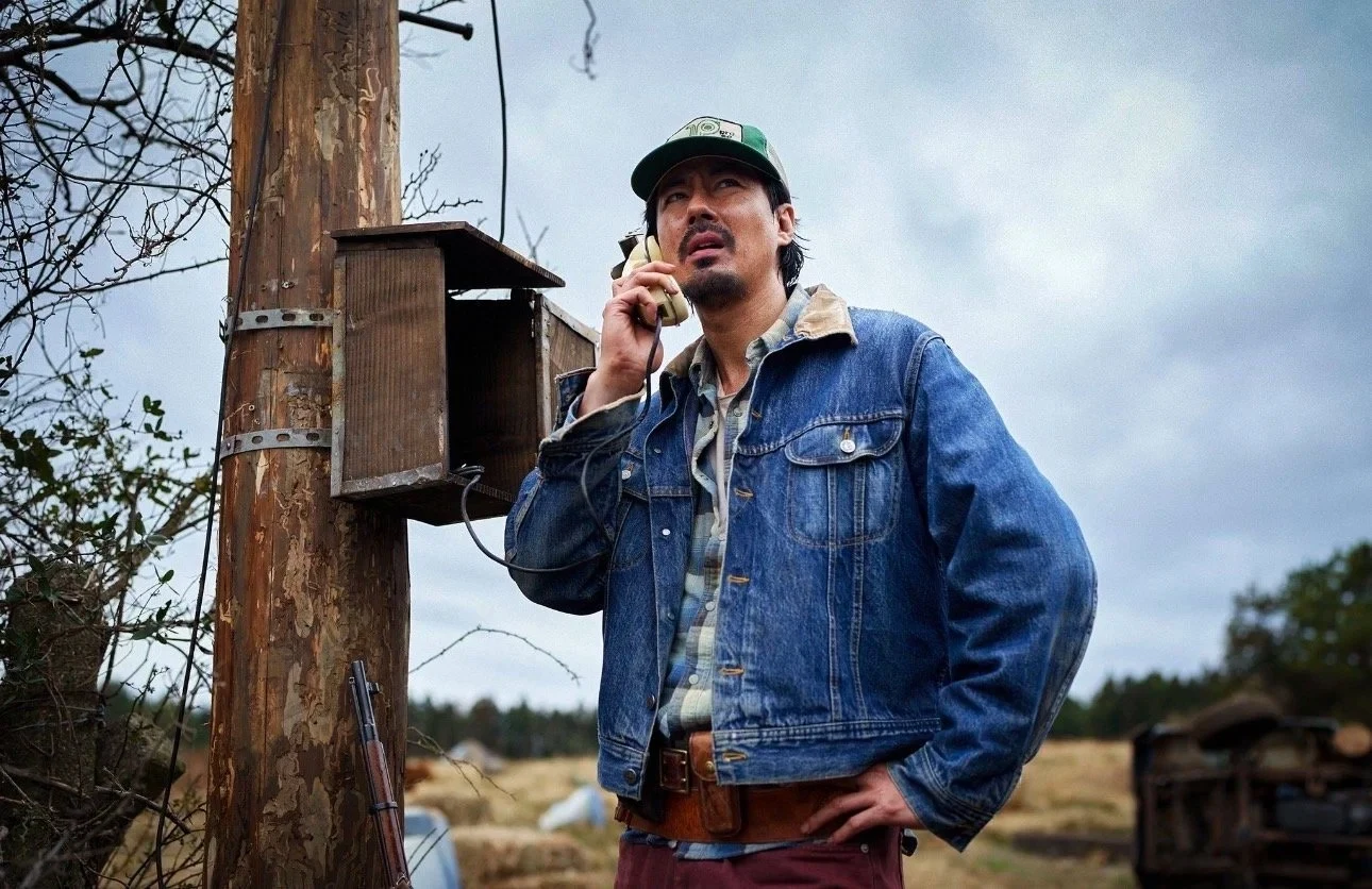 Kadr z filmu "Witajcie w Hope" Mężczyzna w dżinsowej kurtce i czapce z daszkiem przy telefonie na słupie, pole i zachmurzone niebo w tle.