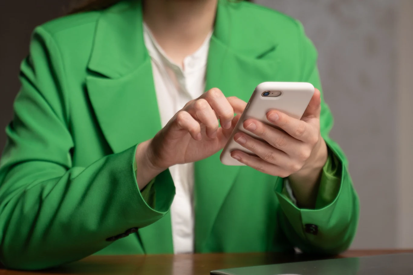 Osoba w zielonej marynarce trzymająca w dłoniach biały smartfon i korzystająca z telefonu.