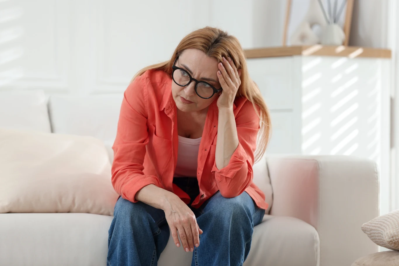 Istnieje kilka sposobów na to, by złagodzić objawy menopauzy. Kobieta w okularach i czerwonej koszuli siedzi na kanapie, podpierając głowę ręką z wyrazem zmęczenia lub zmartwienia.