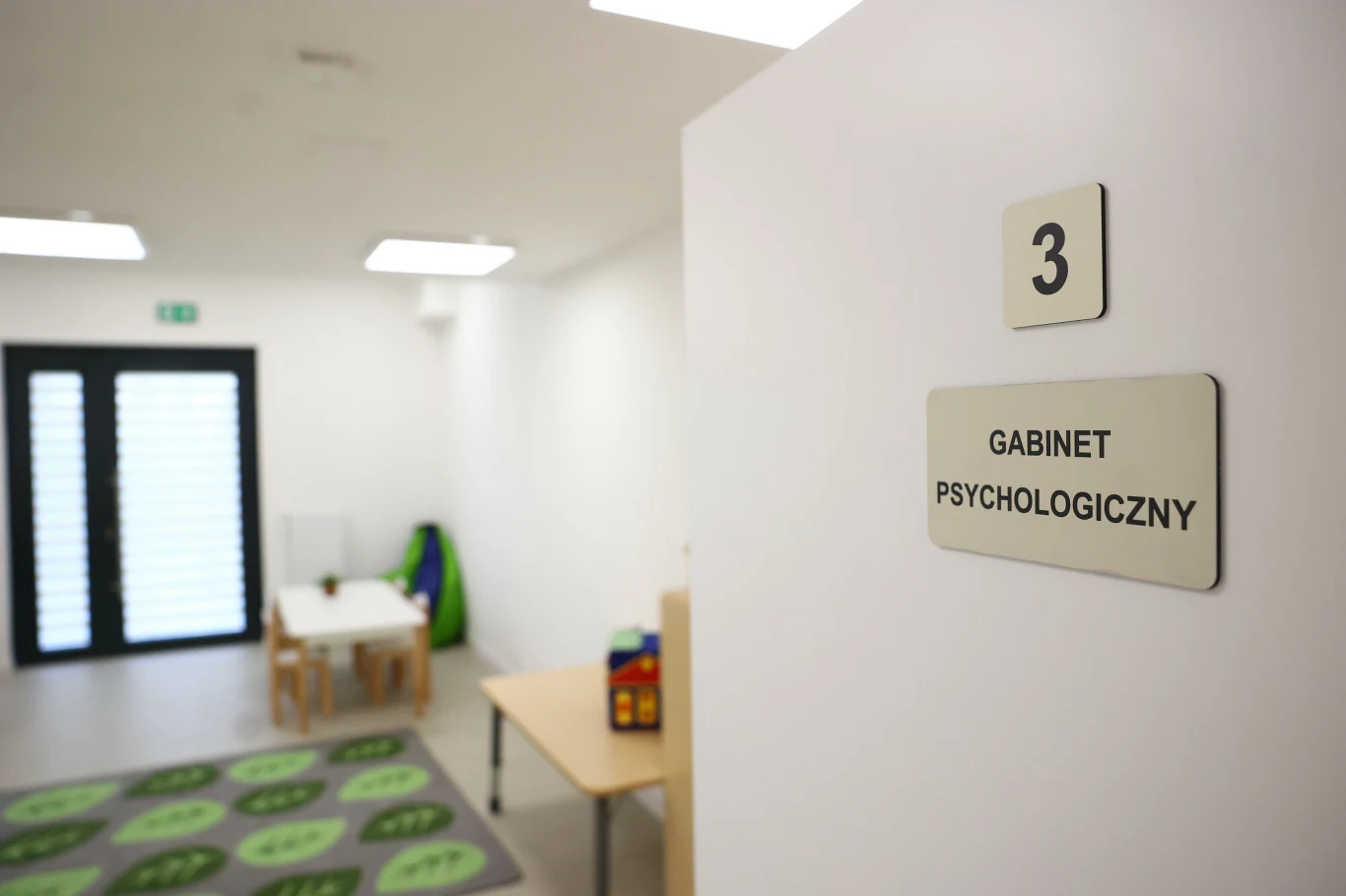 Jasne pomieszczenie z oznaczeniem „Gabinet psychologiczny”, wyposażone w stolik z krzesłami, matę edukacyjną i zabawki, wywołujące atmosferę przyjazną dla dzieci.