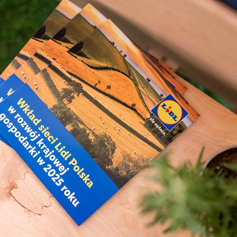 Kolorowe broszury z logo Lidl prezentujące fotografię pól uprawnych i informacje o wkładzie sieci w rozwój krajowej gospodarki w 2025 roku, ułożone na jasnym drewnianym blacie obok doniczki z rośliną.