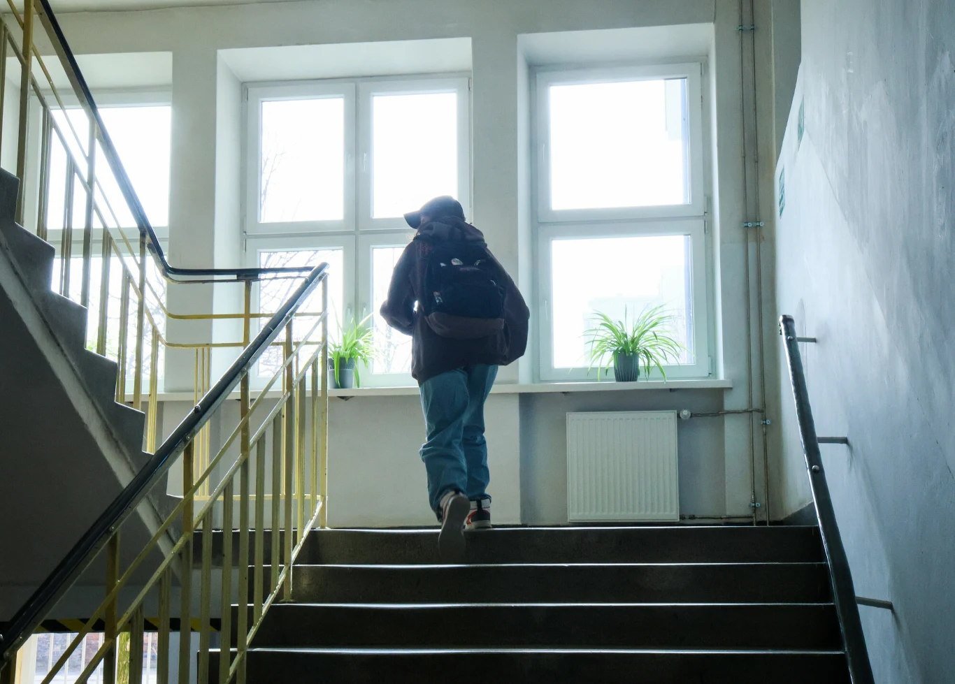 Uczeń z plecakiem idący po schodach szkolnego budynku, jasne światło wpadające przez duże okna oraz dwie zielone rośliny na parapetach.