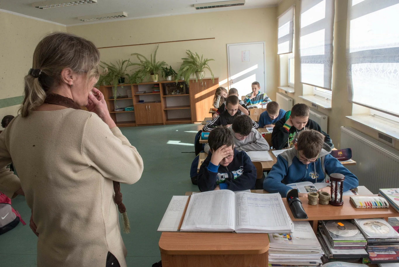 Nauczycielka stojąca przy biurku obserwuje uczniów pochylonych nad zadaniami przy szkolnych ławkach, skupienie dominuje wśród dzieci pracujących indywidualnie.