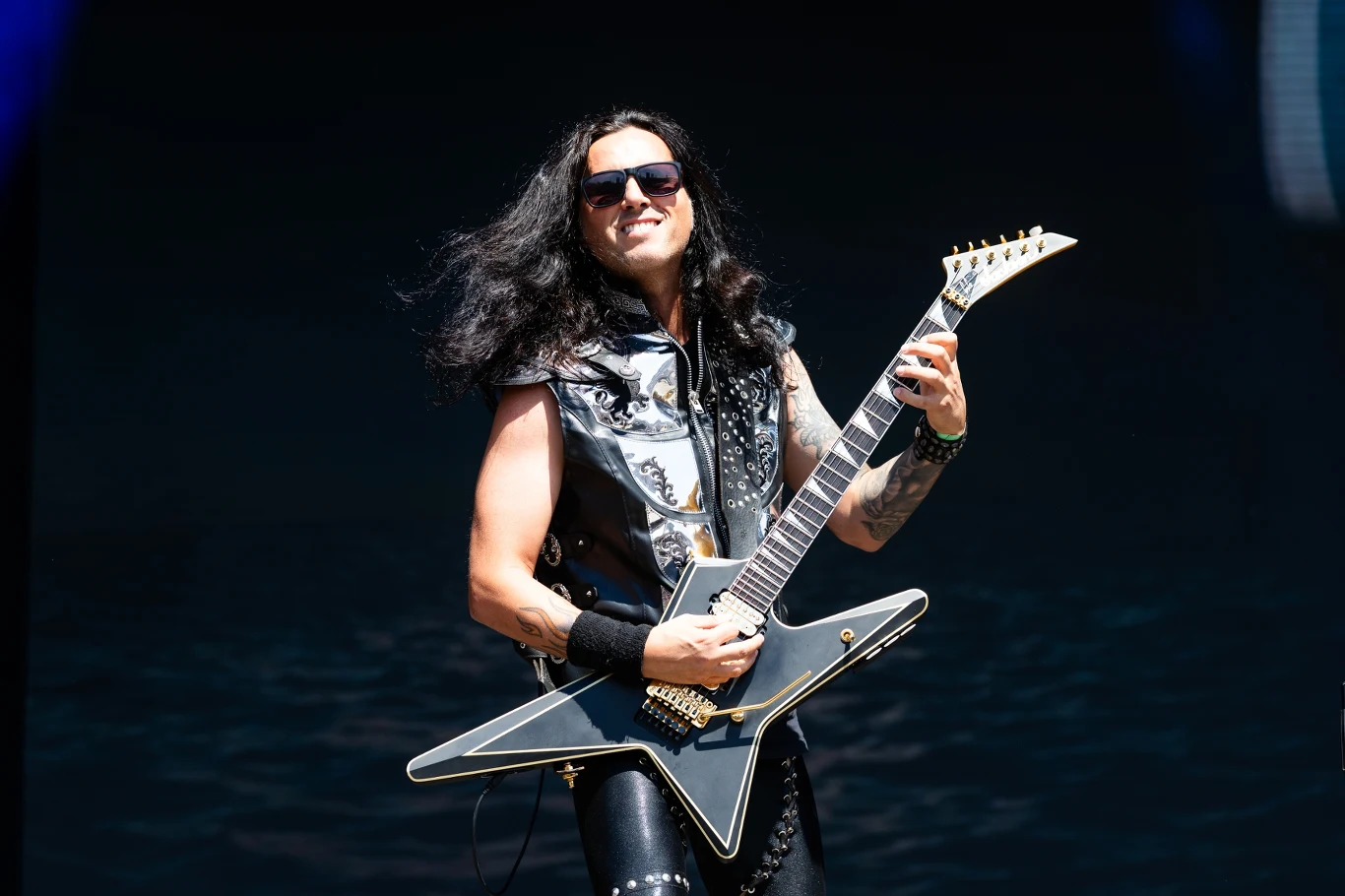 Gus G. jest liderem grupy Firewind Gus G., gitarzysta Firewind, w skórzanej kamizelce gra na czarnej gitarze Flying V na ciemnej scenie.