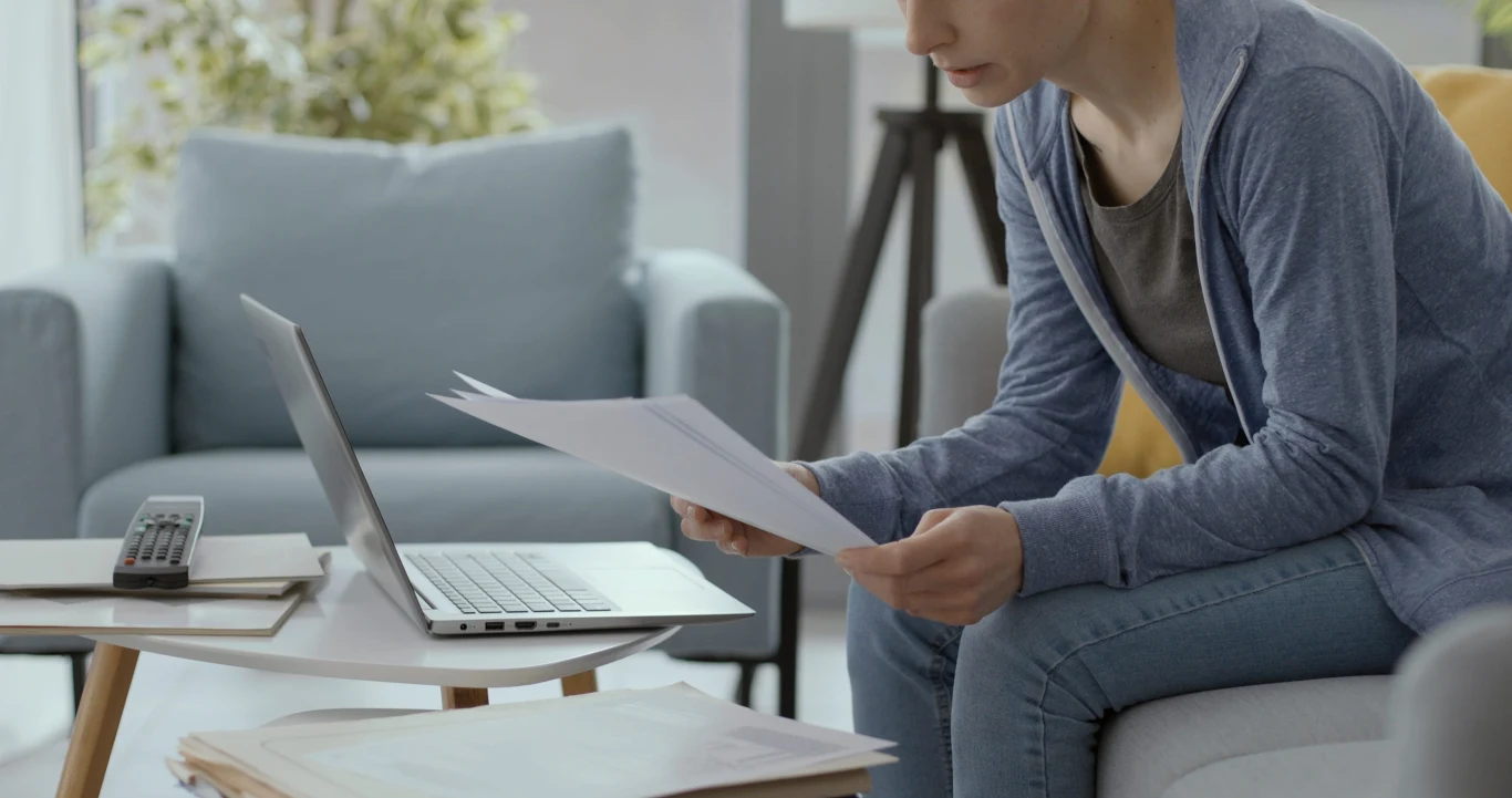 Osoba w niebieskiej bluzie siedzi na sofie, przegląda dokumenty trzymane w rękach, a obok stoi otwarty laptop na stoliku wraz z kilkoma stertami papierów.