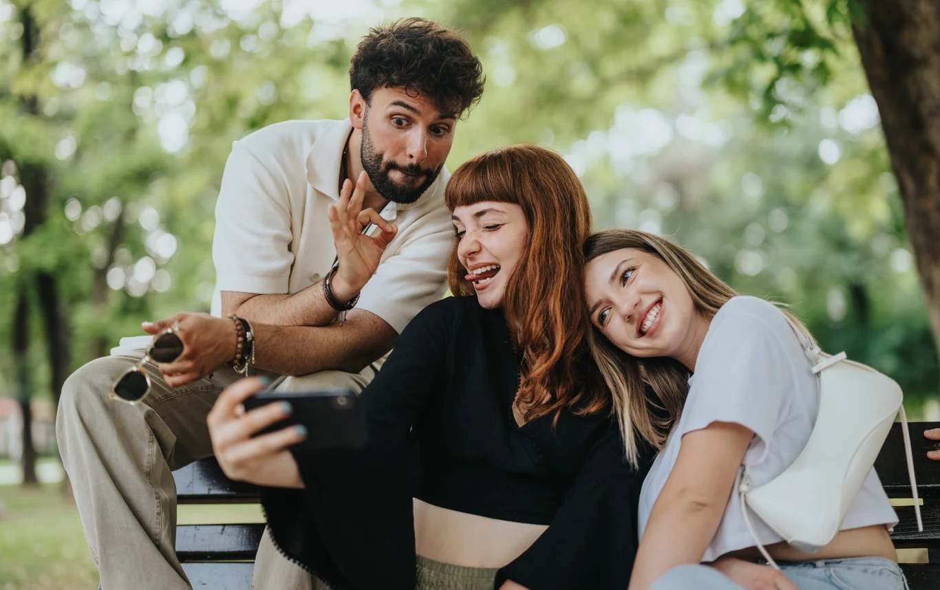 Trzy uśmiechnięte osoby siedzą na ławce, pozując do wspólnego selfie w otoczeniu zielonych drzew.