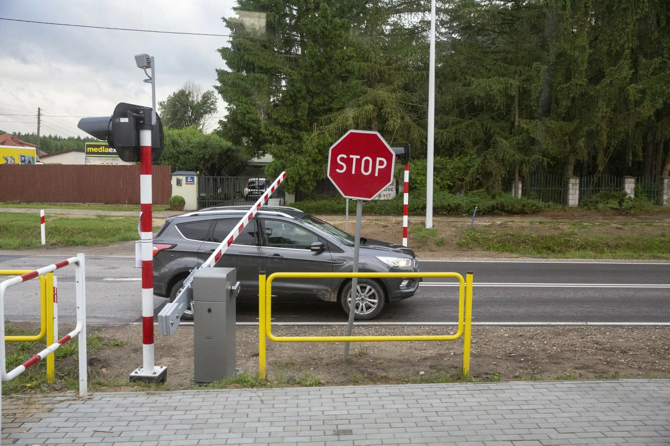 Czarny samochód osobowy stoi przed szlabanem i znakiem stop na tle ulicy otoczonej zielenią, obok system kontroli wjazdu oraz żółte barierki ochronne.