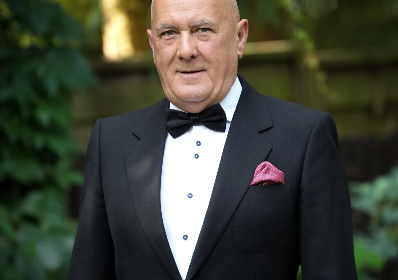 Henryk z Żychlina w "Sanatorium miłości" Elegancki mężczyzna w czarnym smokingu z muchą oraz różową poszetką, stojący na tle zieleni.