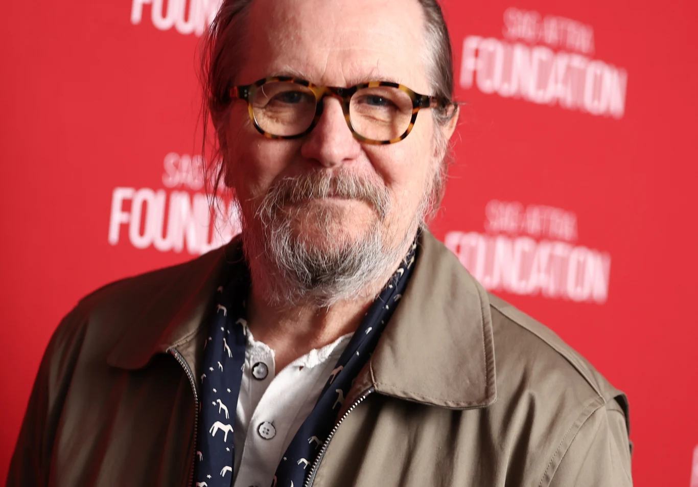 Gary Oldman Gary Oldman w okularach, jasnej koszuli i beżowej kurtce na tle czerwonej ścianki z białymi napisami