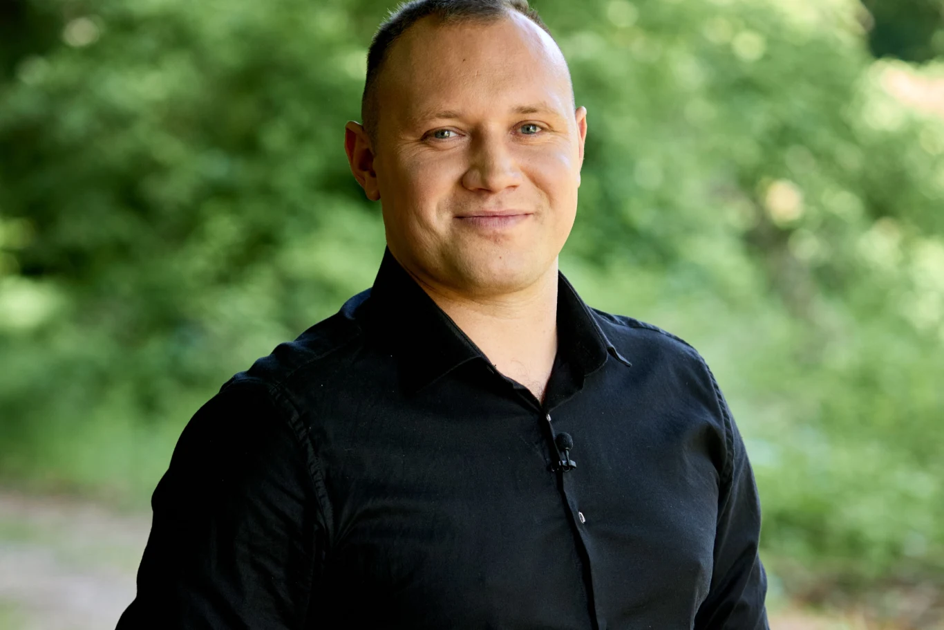 Mężczyzna w czarnej koszuli uśmiecha się, stojąc na tle zieleni drzew.