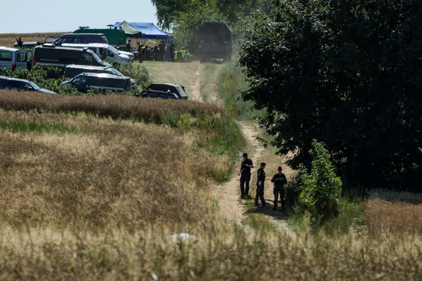 Grupa funkcjonariuszy stoi na polnej drodze wśród wysokich traw, w tle widoczne są zaparkowane pojazdy służbowe oraz namioty rozstawione na skraju pola.
