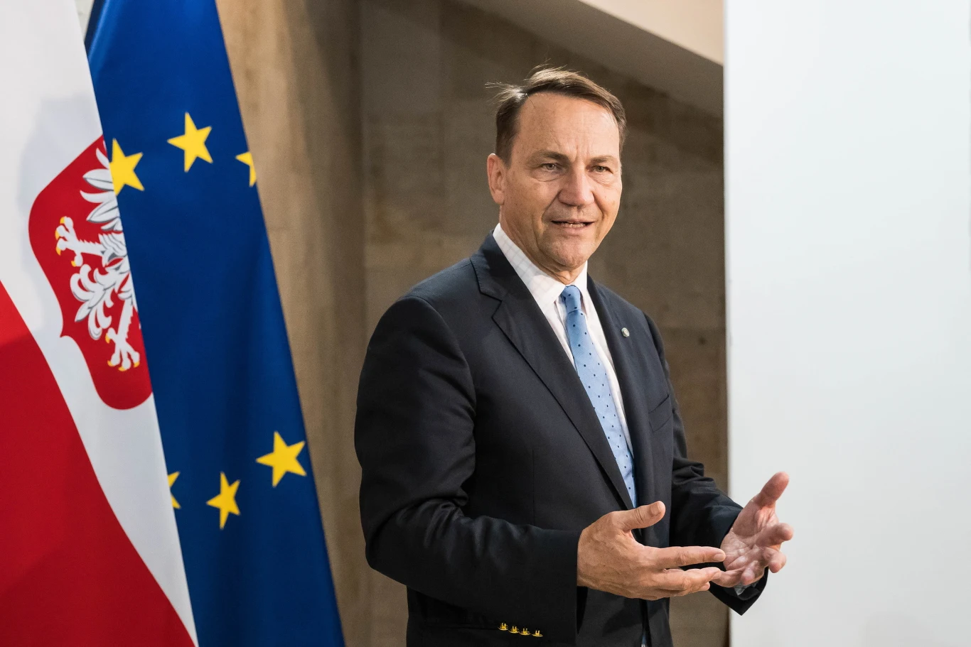 Szef MSZ Radosław Sikorski podczas wizyty w Kijowie Radosław Sikorski w garniturze i niebieskim krawacie podczas wystąpienia w Kijowie, przy flagach Polski i UE.