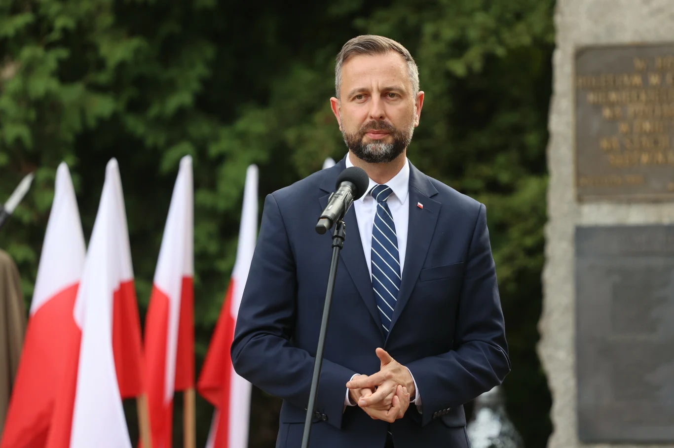 Szef MON Władysław Kosiniak-Kamysz Władysław Kosiniak-Kamysz przy mikrofonie, w garniturze, na tle polskich flag i fragmentu pomnika.