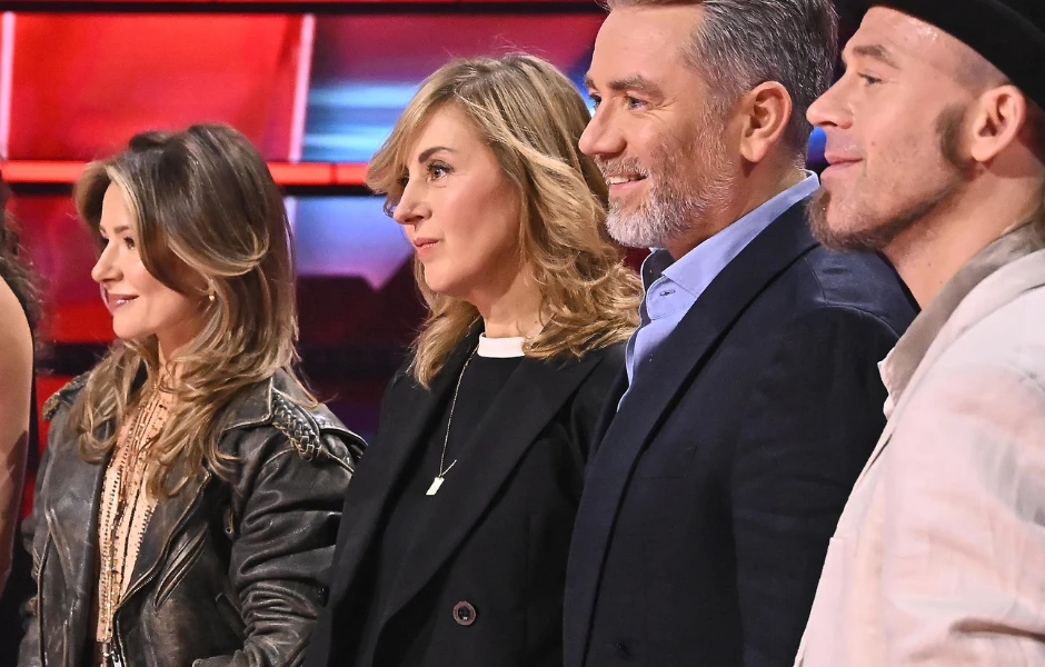 "The Voice of Poland" Czworo dorosłych osób w eleganckich i casualowych stylizacjach stoi obok siebie, skupiając uwagę w jednym kierunku, na tle intensywnych kolorów i świateł.