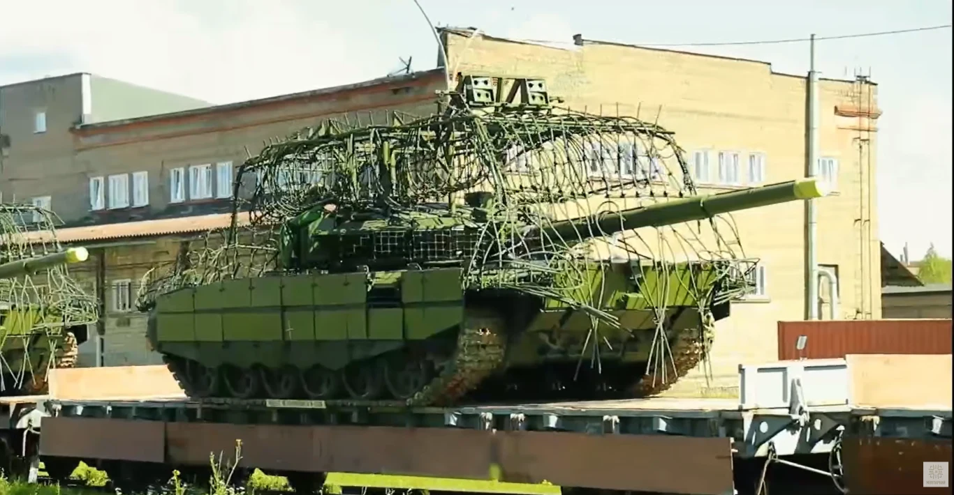 T-90M i T-72B3M dla rosyjskiej armii. Czołgi przygotowane na zagrożenie z powietrza Czołg T-90M z siatką ochronną na platformie kolejowej, w tle budynek przemysłowy.