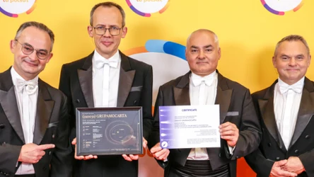 Czterech elegancko ubranych mężczyzn we frakach, jeden trzyma czarną tablicę z podpisem, drugi certyfikat, wszyscy na tle żółtej, kolorowej ścianki.