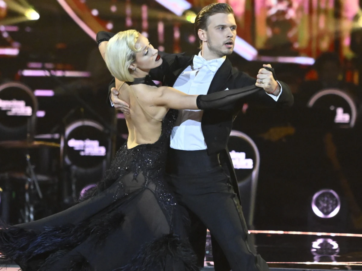 Para elegancko ubranych tancerzy w dynamicznym tańcu standardowym, kobieta w czarnej sukni z odkrytymi plecami, mężczyzna w fraku, oboje skupieni na ruchu i muzyce, scena oświetlona reflektorami.