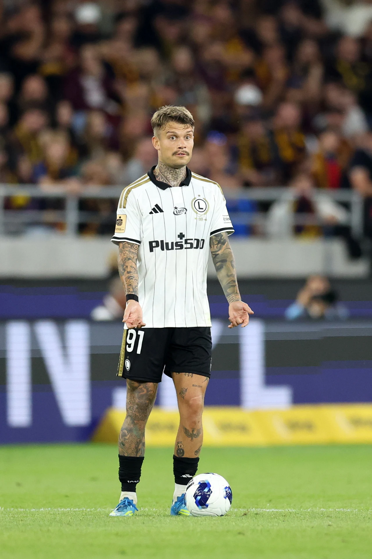 Kamil Piątkowski Piłkarz w białej koszulce z numerem 91 i logotypem Legii Warszawa stoi na murawie z piłką przy nodze, koncentrując się przed wykonaniem stałego fragmentu gry.