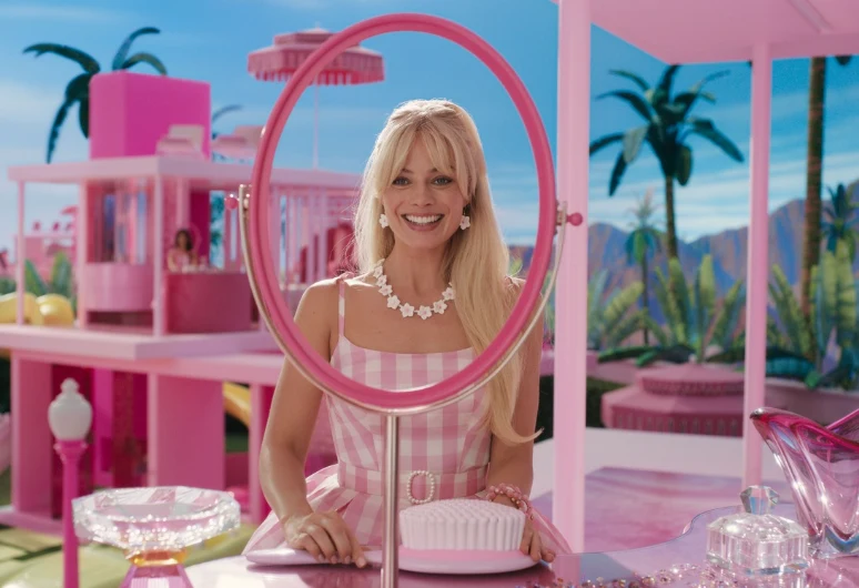 Kadr z filmu "Barbie" Margot Robbie jako Barbie w różowej sukience siedzi przed toaletką w różowym wnętrzu z palmami i pastelowymi budynkami.