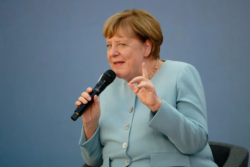 Angela Merkel coraz częściej komentuje bieżącą politykę Kobieta w jasnoniebieskiej marynarce siedzi na krześle, trzyma mikrofon i unosi palec wskazujący, jakby podkreślała ważny punkt podczas wypowiedzi.