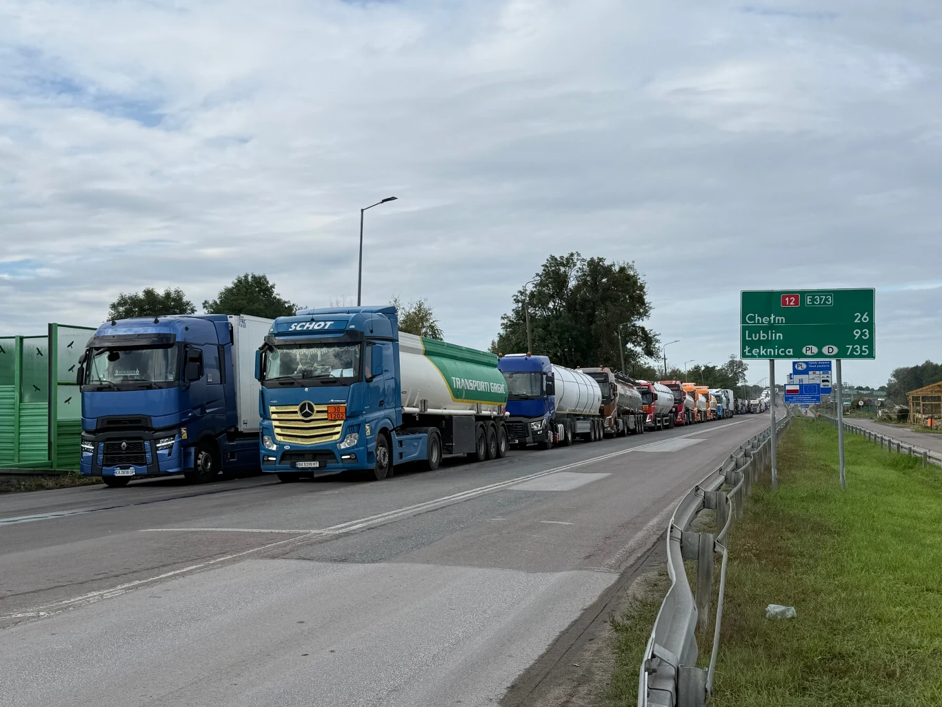 Ciężarówki czekające w kolejce na przejściu granicznym Szereg ciężarówek ustawionych w długim korku przy głównej drodze obok zielonego drogowskazu z informacją o odległościach do Chełma, Lublina i Leśnicy.