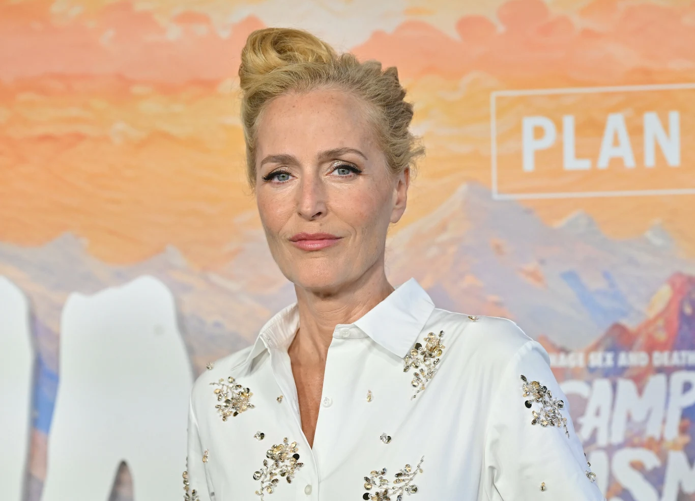 Gillian Anderson Gillian Anderson w białej koszuli z aplikacjami, blond włosy w kok, na tle malarskiej scenografii z motywem górskim.