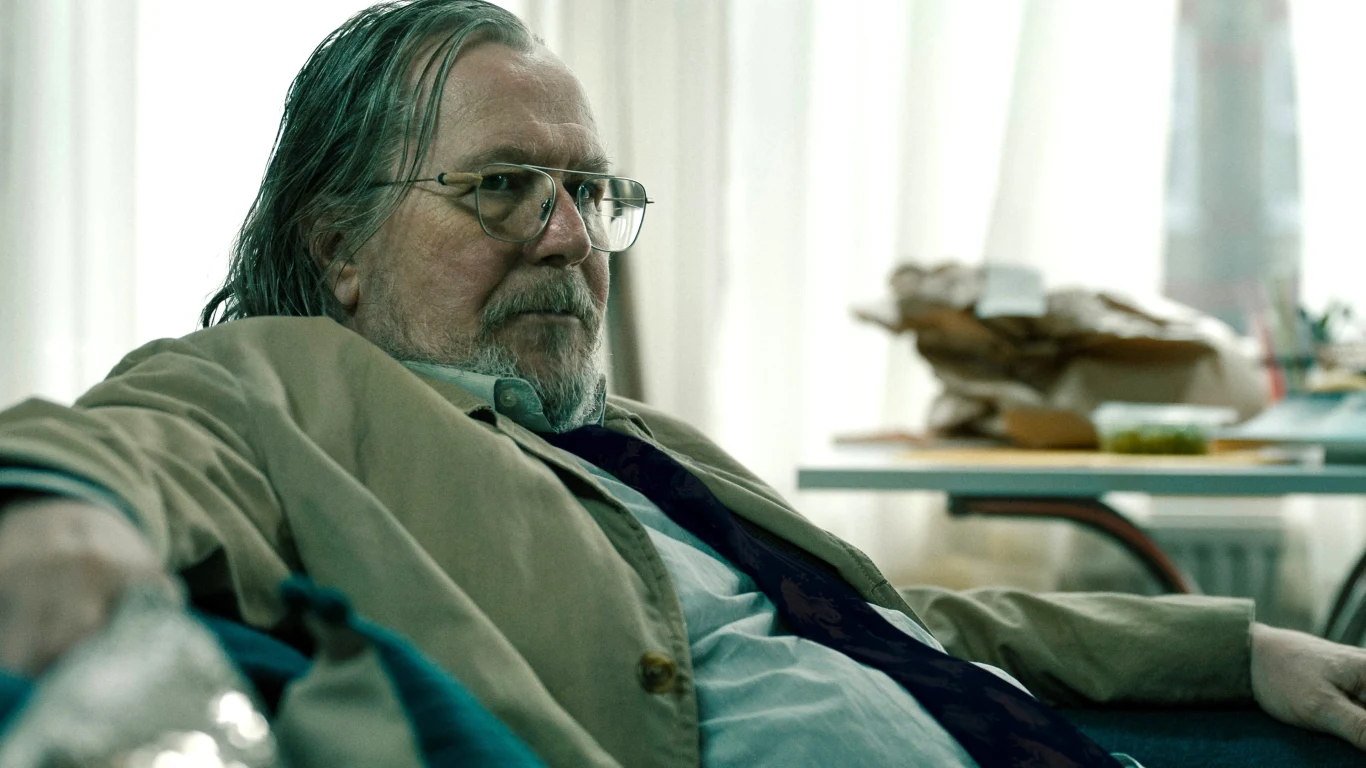 Gary Oldman w serialu "Kulawe konie" Gary Oldman jako Jackson Lamb w serialu 'Kulawe konie', starszy mężczyzna w okularach siedzi na kanapie