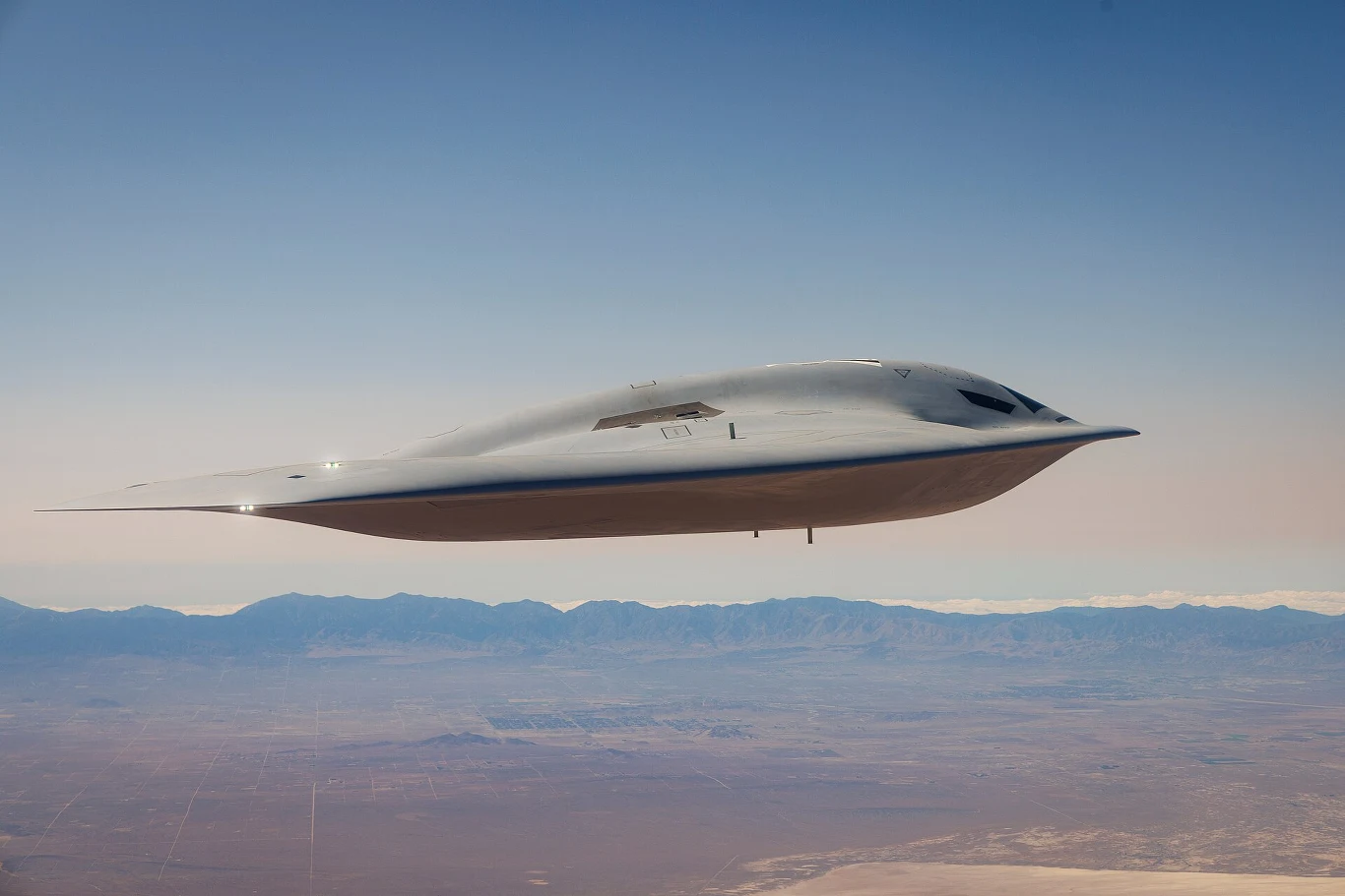 B-21 Raider podczas lotów testowych. Nowoczesny bombowiec strategiczny o opływowym, futurystycznym kształcie leci nad pustynnym, górzystym terenem.