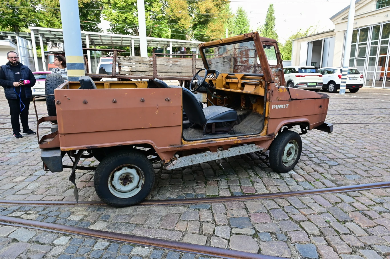 Stary, mocno zużyty, pozbawiony dachu samochód terenowy w brązowym kolorze stoi zaparkowany na brukowanej powierzchni w otoczeniu torów tramwajowych, z widocznymi prowadzącymi rozmowę ludźmi w tle.
