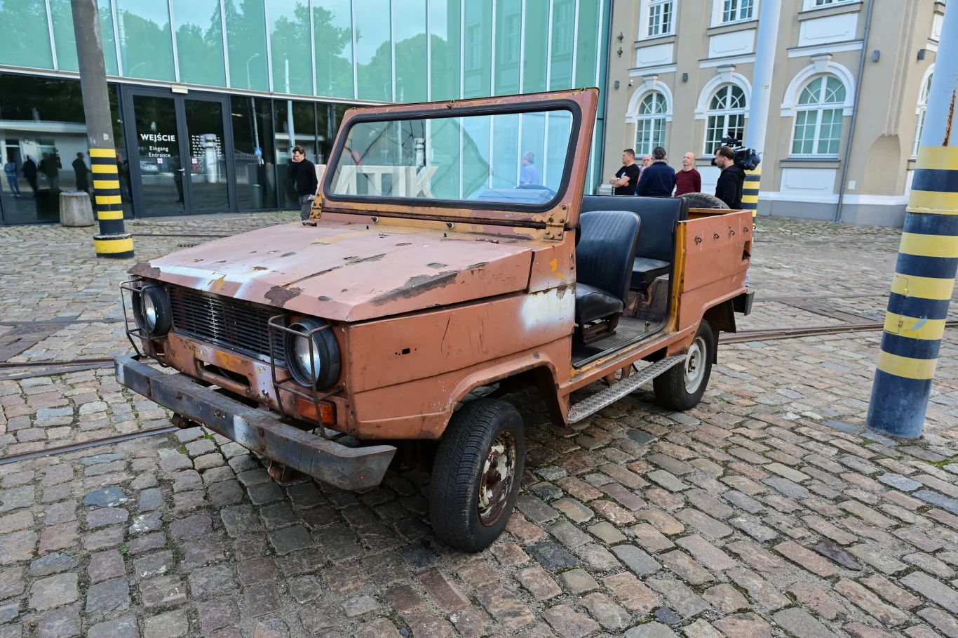 Zardzewiały, pozbawiony drzwi terenowy samochód w kolorze brązowym ustawiony na brukowanym placu przed nowoczesnym budynkiem, w tle grupa osób rozmawiających przy wejściu.