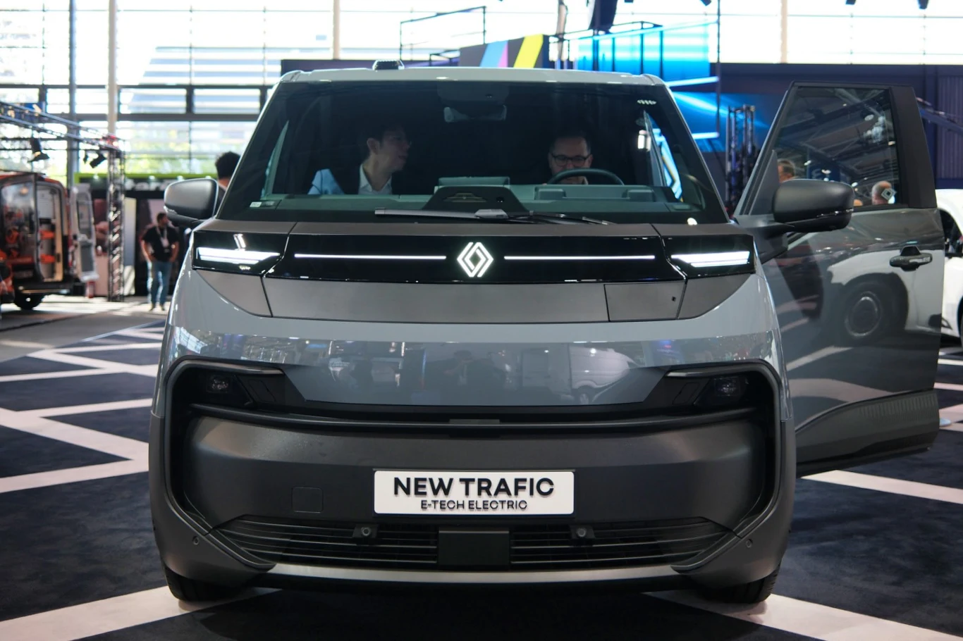 Nowoczesny van Renault New Trafic E-Tech Electric ustawiony na ekspozycji targowej z otwartymi drzwiami kierowcy, wyraźnie widoczny przedni grill i charakterystyczne oświetlenie LED.