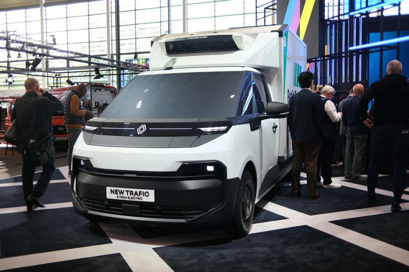 Nowoczesny biały samochód dostawczy Renault New Trafic z zabudową chłodniczą, otoczony przez grupę osób podczas prezentacji na targach motoryzacyjnych.