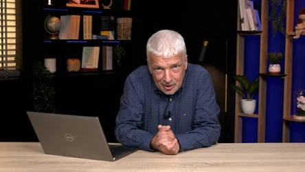 Starszy mężczyzna z siwymi włosami pochyla się nad biurkiem z laptopem, w tle półki z książkami, rośliny i akcesoria biurowe.