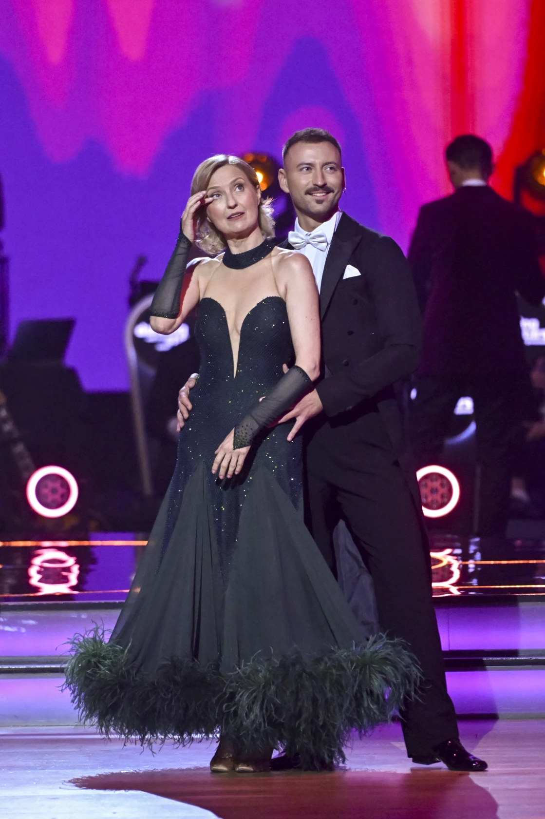 Para tancerzy w eleganckich strojach w pozie tanecznej na kolorowo oświetlonej scenie, kobieta w długiej, ciemnej sukni z piórami i długimi rękawiczkami, mężczyzna w klasycznym smokingu.