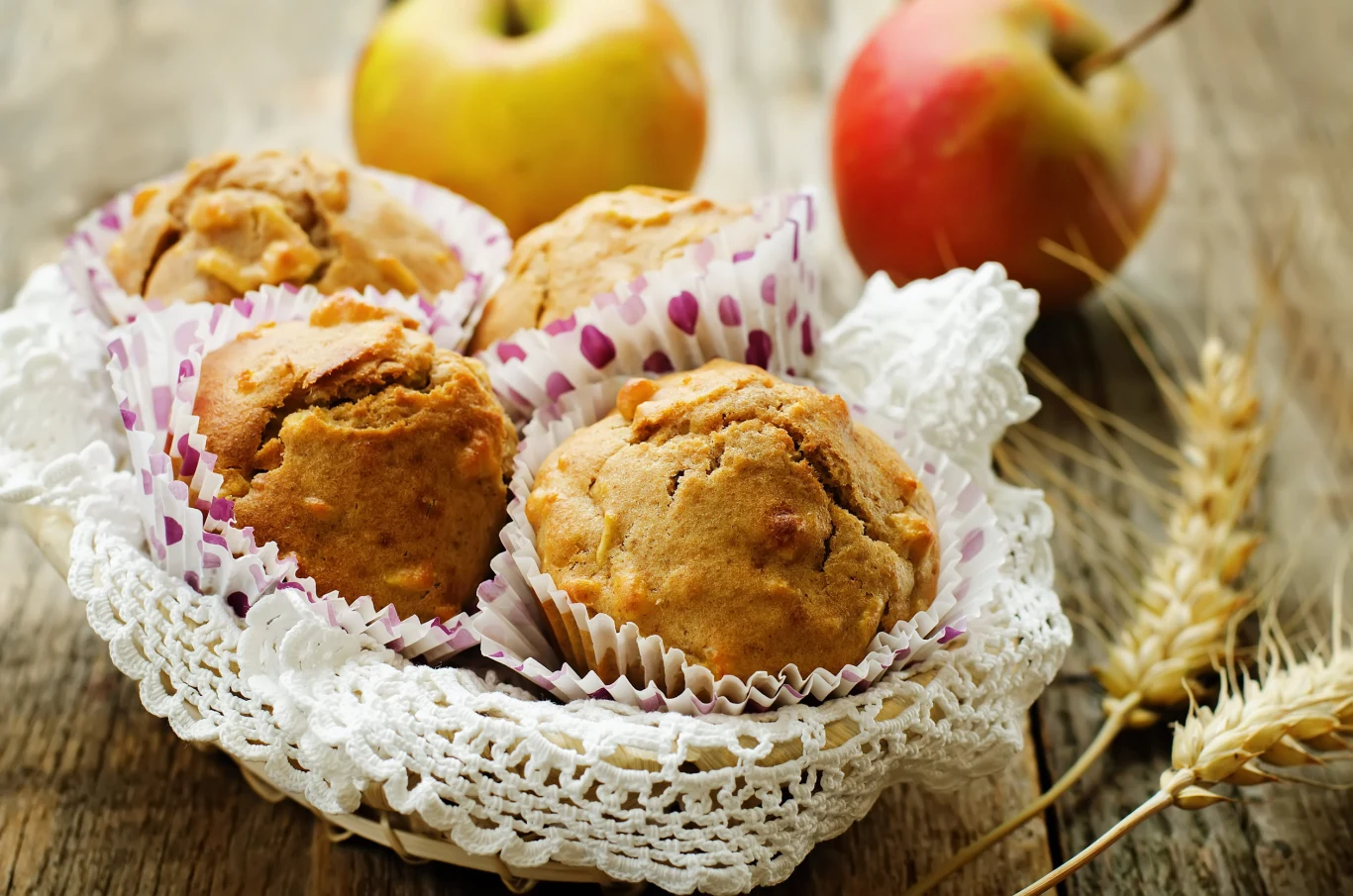 Muffinki jabłkowe to puszysty, wilgotny i miękki smakołyk do popołudniowej kawy Koszyk z domowymi muffinkami jabłkowymi w papilotkach, koronkowa serwetka, w tle jabłka i kłosy zboża.