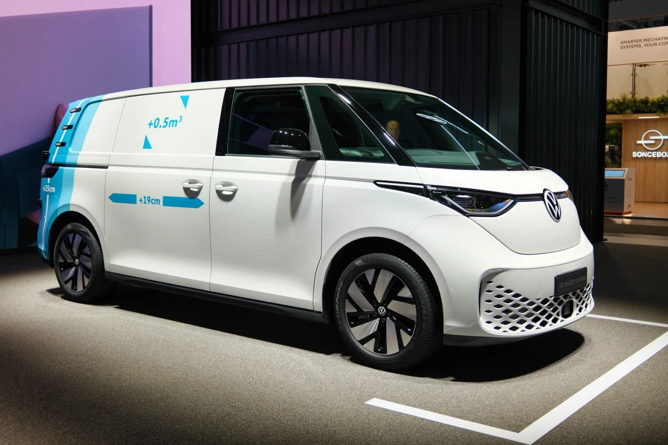 Nowoczesny biały van z czarnymi detalami i niebieskimi grafikami na bocznych drzwiach, charakterystyczny znak Volkswagena z przodu pojazdu, duże felgi i dynamiczna sylwetka podkreślające użytkowy charakter samochodu.
