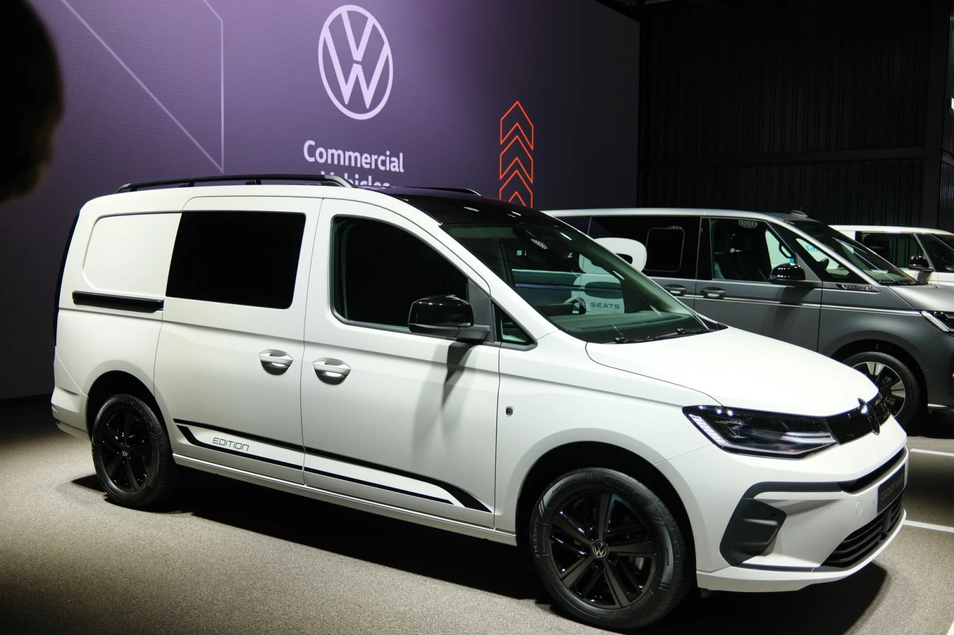 Biały van Volkswagen Caddy z czarnymi felgami i przyciemnianymi szybami prezentowany na stoisku wystawowym, w tle widoczne logo marki oraz inny samochód tej samej firmy.