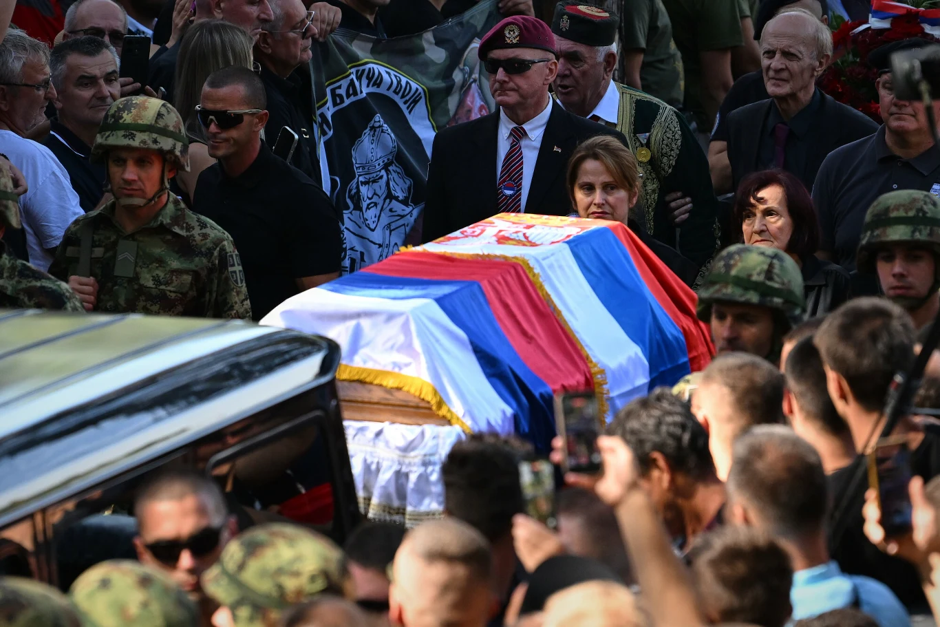 Trumna przykryta serbską flagą niesiona przez tłum żołnierzy i cywilów podczas ceremonii pogrzebowej, otoczona przez liczną grupę ludzi w skupieniu.