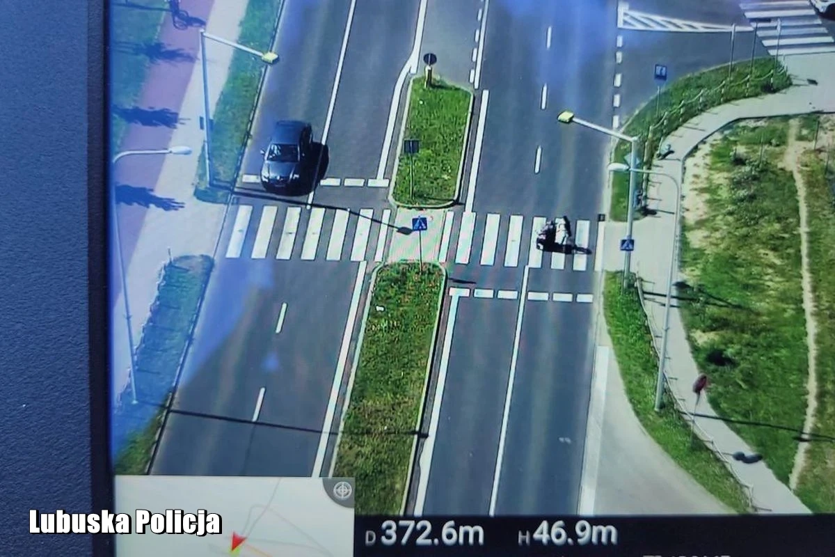 Z drona okolice przejścia widać jak na dłoni Samochód osobowy zbliżający się do przejścia dla pieszych na skrzyżowaniu, przez które przechodzi dwójka pieszych.